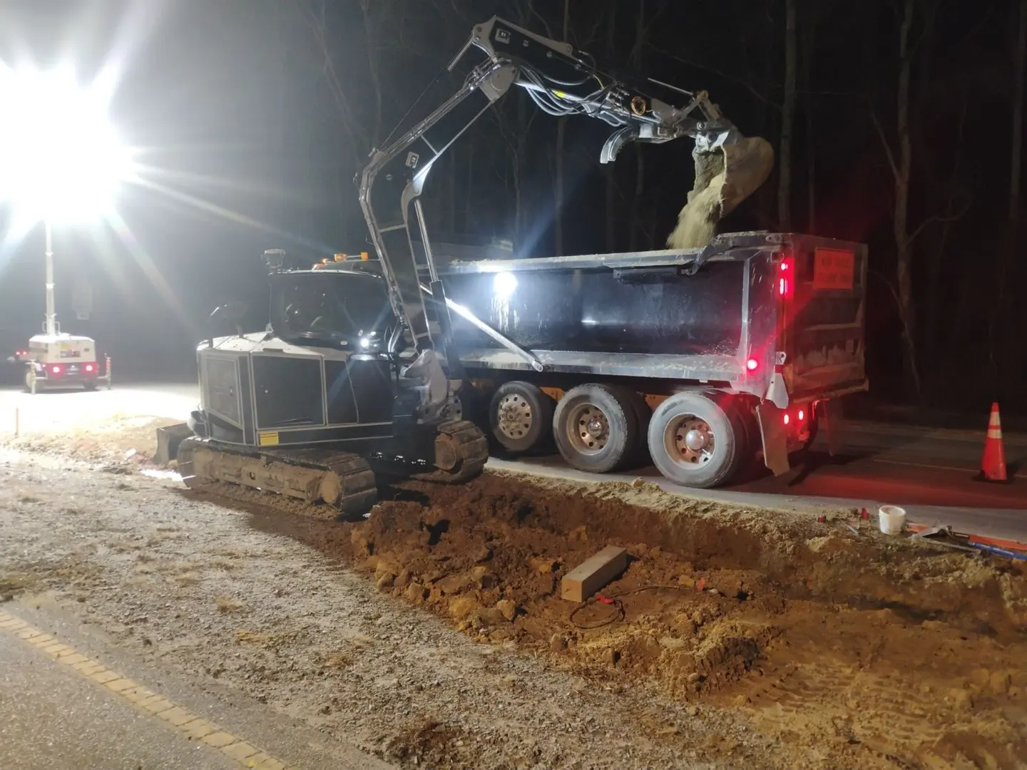 An excavator loads soil into a dump truck at a lighted nighttime work area.